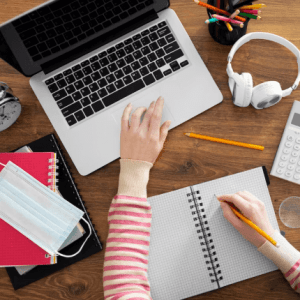 An overhead shot of a person's hands, one on a laptop and the other writing in a notebook, surrounded by study materials and a face mask.