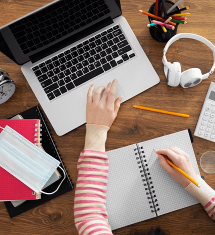 An overhead shot of a person's hands, one on a laptop and the other writing in a notebook, surrounded by study materials and a face mask.