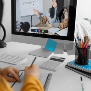 A student's hands taking notes from a computer screen displaying "LESSON 1" with a teacher explaining, in a home study setting.