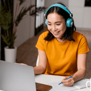 A smiling woman in a yellow shirt wearing headphones works at her laptop with notes and a pen in hand, indoors.