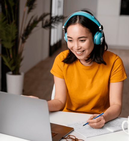 A smiling woman in a yellow shirt wearing headphones works at her laptop with notes and a pen in hand, indoors.