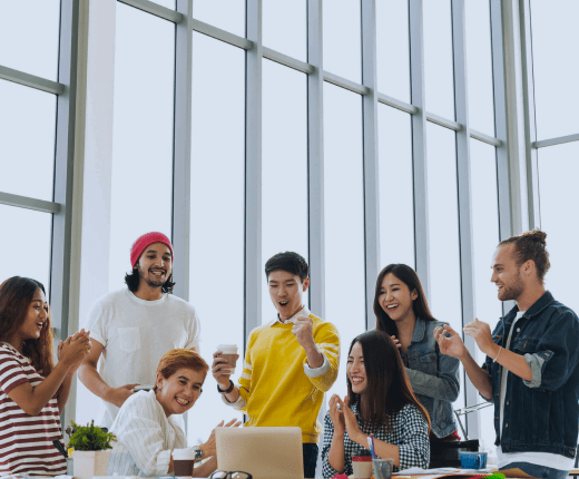 Modern office with seven professionals cheering around a table with laptops.