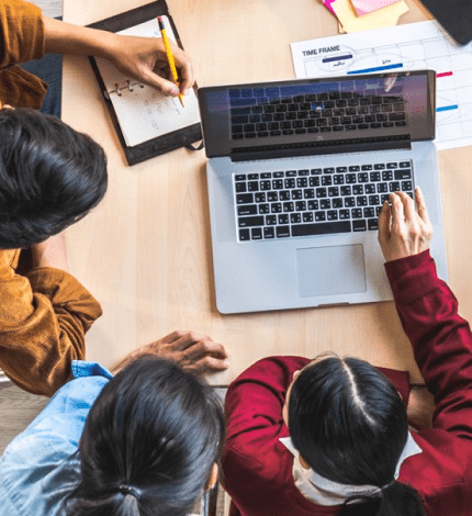 Conference room with a large table and 3 individuals collaborating in front of a laptop.
