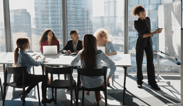 Employees in a modern office workspace with a desktop computer and stationery on a clean desk in front of a white board collaborating.