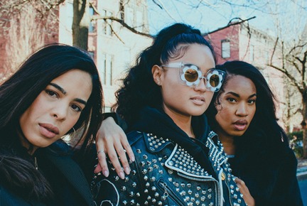 Three confident women posing on a city street, with one in the center wearing sunglasses and a studded jacket.